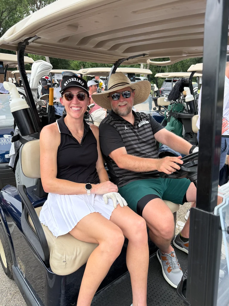 Two alumni smiling from a golf cart during the outing