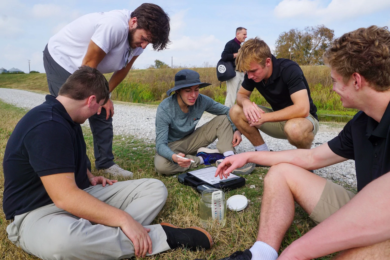 Cistercian students gather beside a pond to review water testing equipment and samples during an outdoor science lesson.
