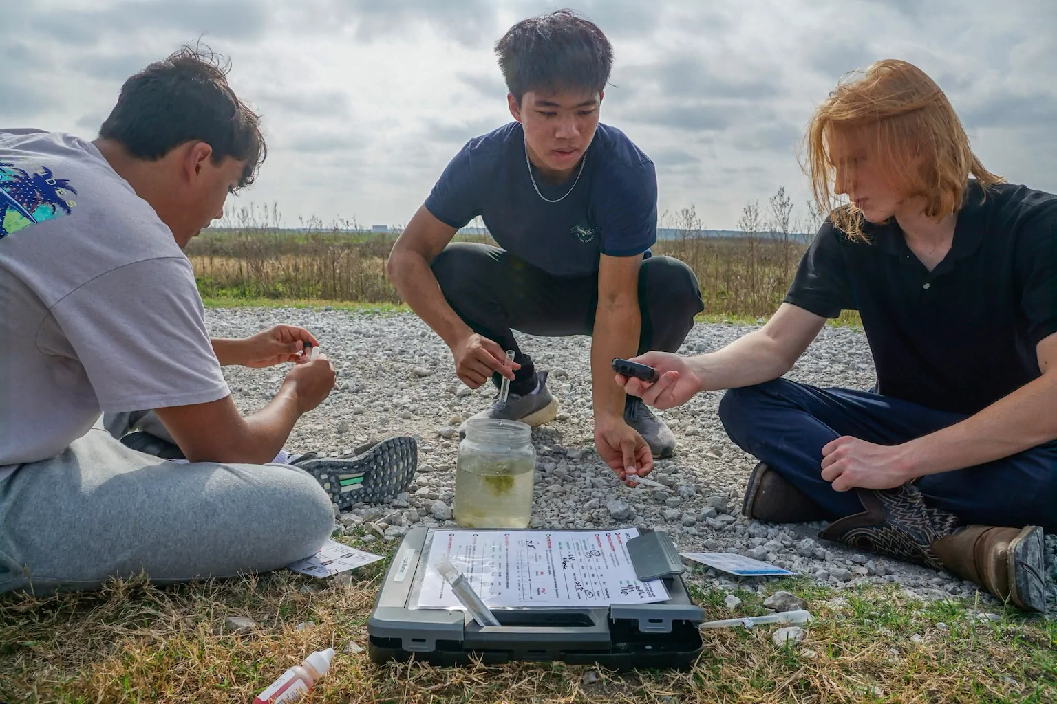 Students analyze a water sample with testing materials during a field lesson near a pond.