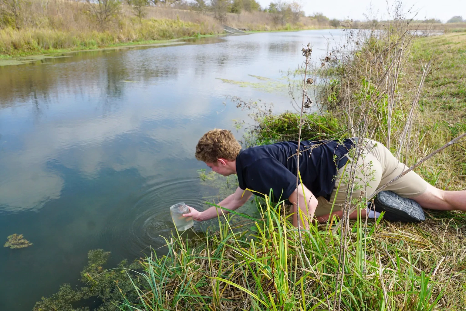 A student collects pond water in a jar for environmental science testing.